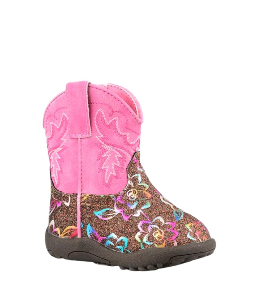 A toddler's western-style boot featuring a bright pink faux-suede shaft with embroidered stitching, a glittery brown footbed decorated with a colorful floral pattern, and a dark brown sole.