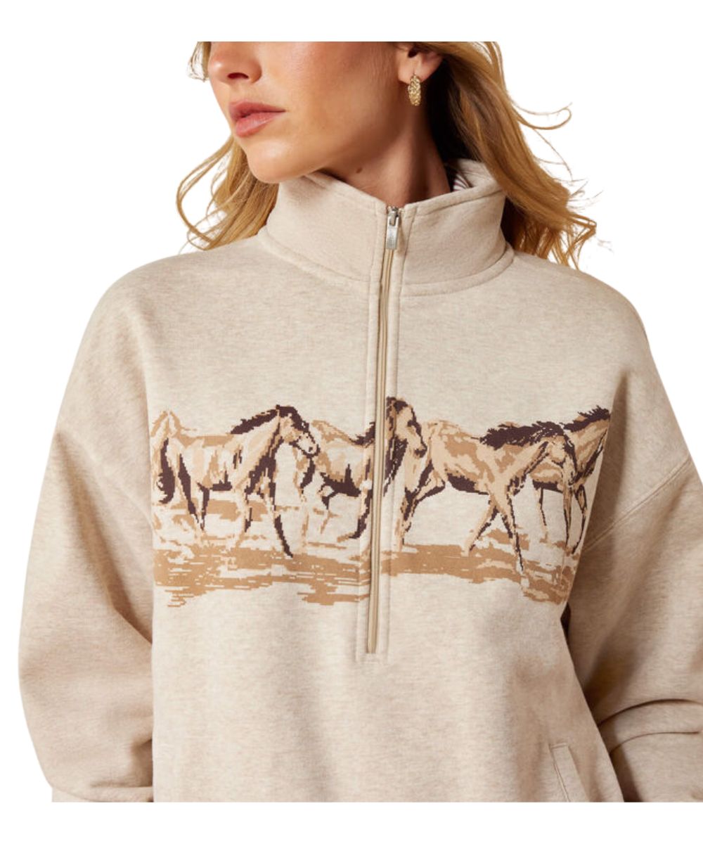 A woman wearing a light beige heathered quarter-zip sweatshirt with a graphic print of running horses across the chest.