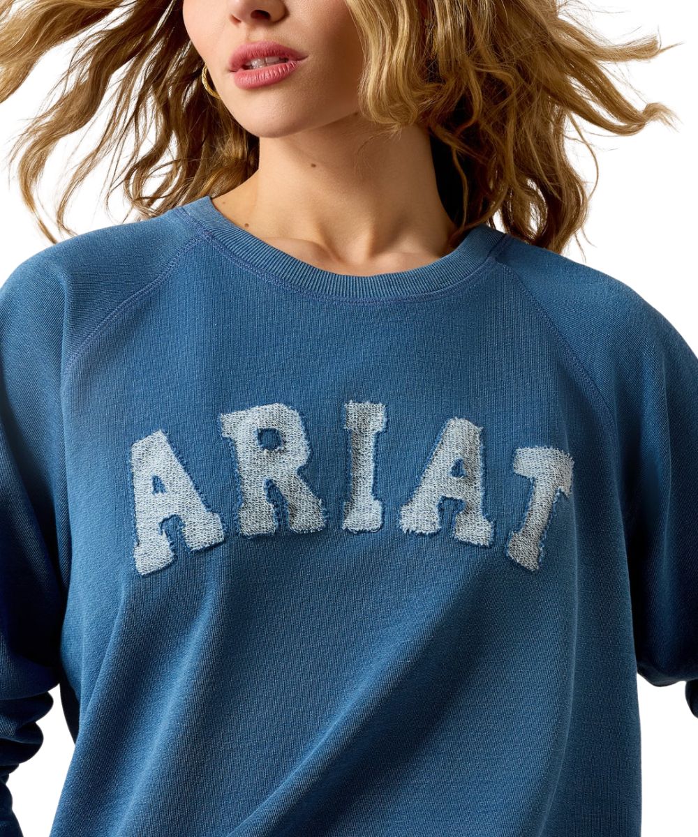 A close-up of a woman wearing a blue crewneck sweatshirt with 'ARIAT' embroidered in large white letters across the chest.