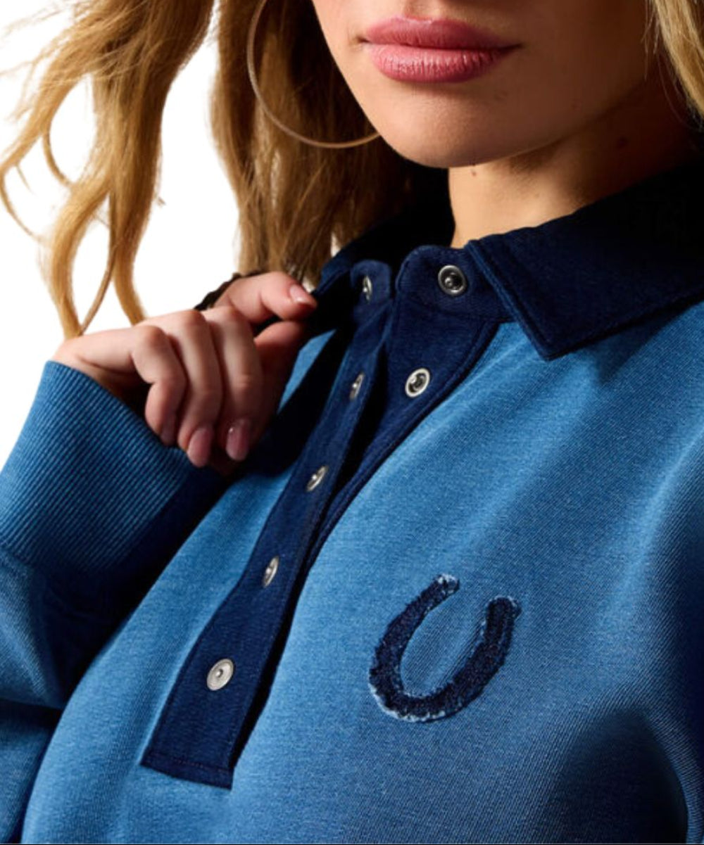 Close-up of a woman wearing a blue knit polo-style top with a dark navy collar and placket, silver buttons, and a dark embroidered horseshoe logo on the chest.