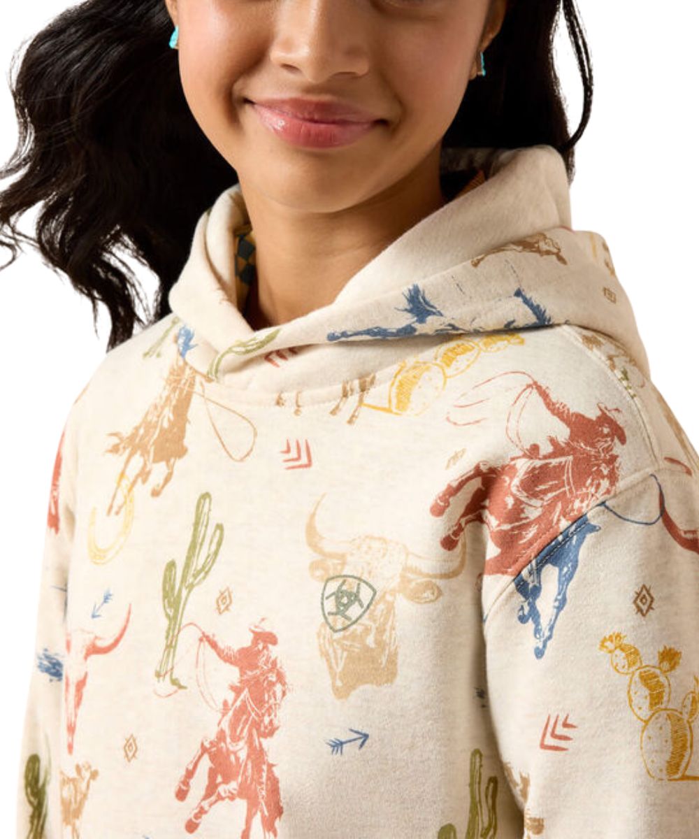 A close-up of a person wearing a cream-colored hooded sweatshirt with a colorful western-themed print of cowboy boots, cacti, and rodeo animals, against a white background.