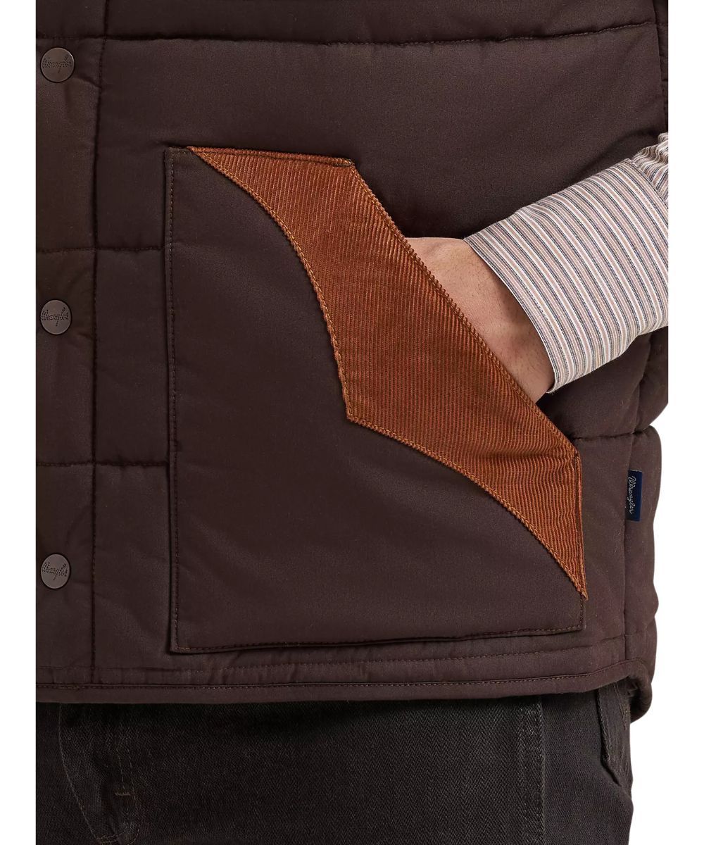 Close-up of a dark brown quilted puffer vest with a contrasting brown corduroy pocket panel and a striped shirt sleeve tucked into the pocket.