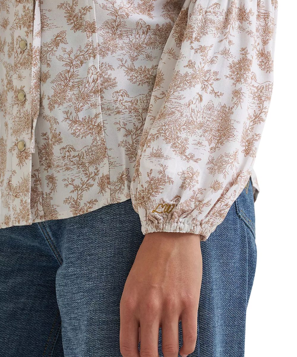 A close-up of a person wearing a cream-colored blouse with a brown toile floral pattern and blue jeans. The blouse features gathered, balloon-style sleeves with elastic cuffs, and a button-down front.