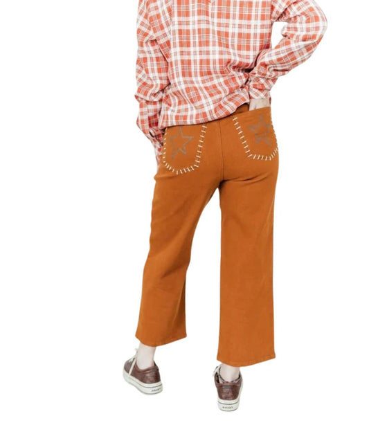 A person shown from the back wearing rust-orange cropped pants with embroidered star details on the back pockets, an orange-and-white plaid shirt, and brown sneakers, against a plain white background.