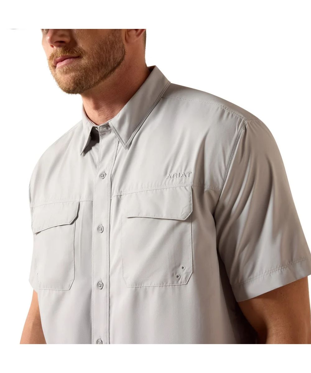 A close-up of a man wearing a light gray short-sleeve button-up shirt with two chest pockets and a subtle 'Ariat' logo on the left chest, set against a plain white background.