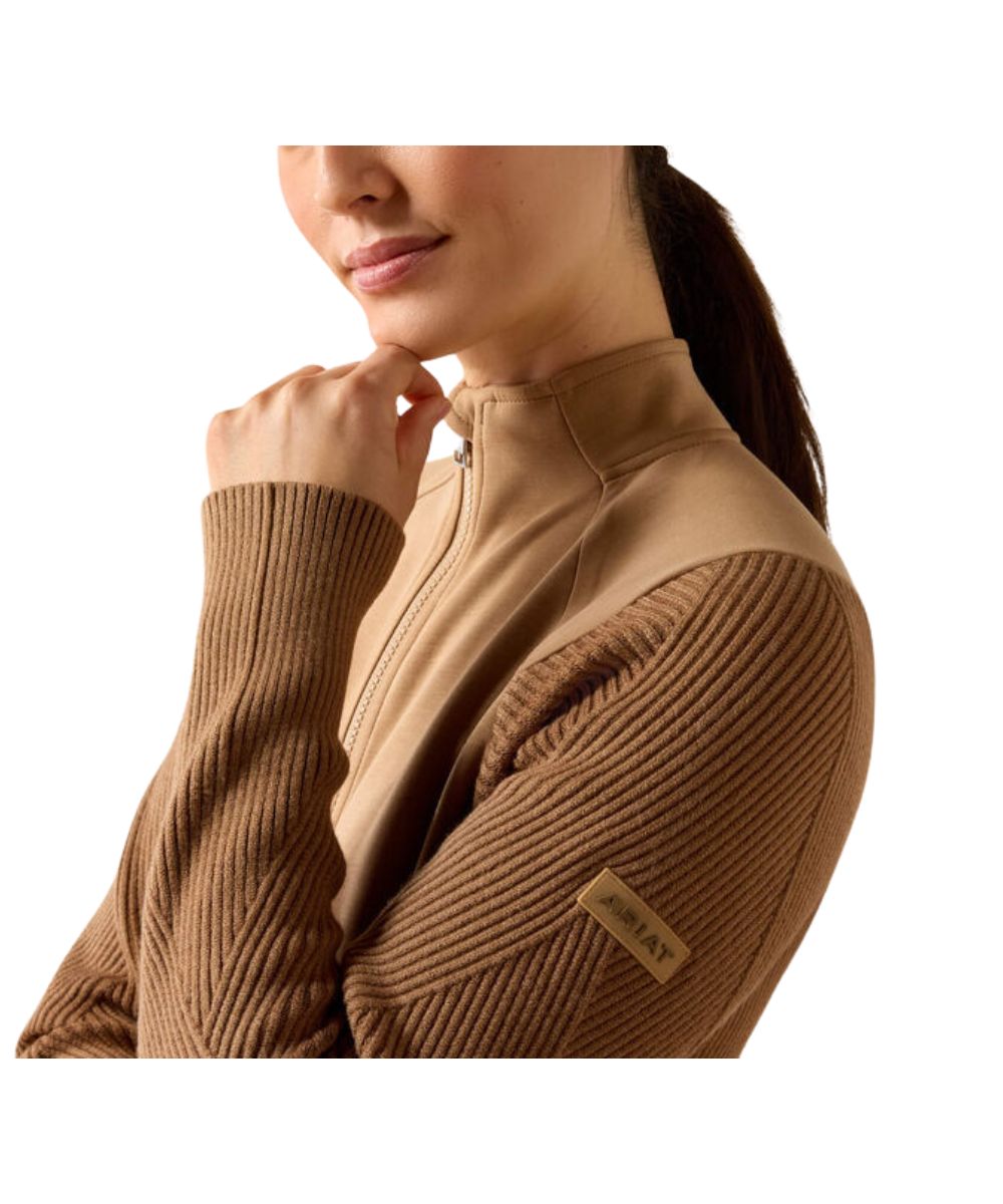 A close-up of a woman wearing a tan and brown quarter-zip fleece jacket with ribbed knit sleeves, featuring a small 'ARIAT' brand patch on the sleeve, set against a plain white background.