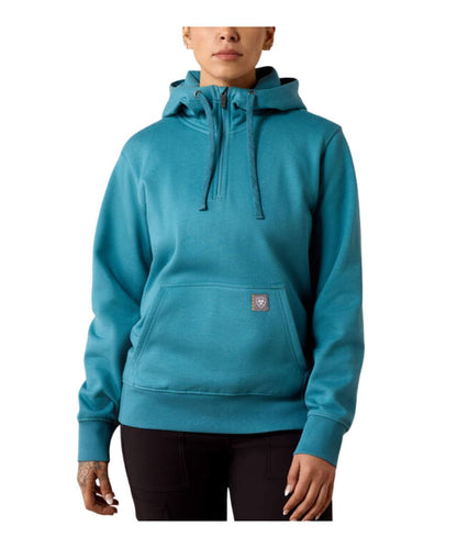 A woman wearing a teal blue pullover hoodie with a half-zip neckline, drawstring hood, and a small shield logo patch on the front pocket, paired with black pants against a plain white background.