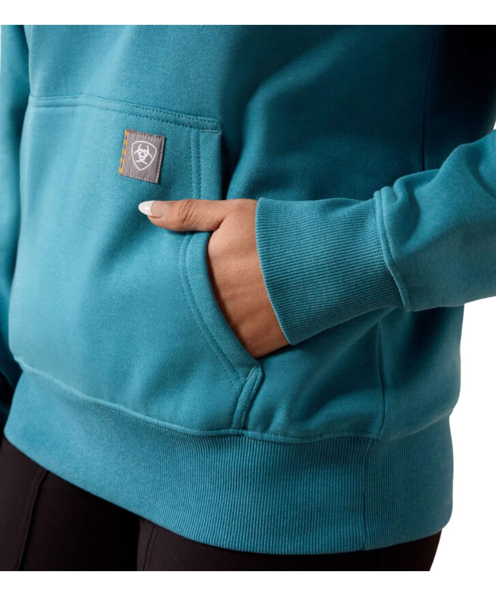 A close-up of a person wearing a teal blue fleece hoodie with a front pocket and a small square brand patch on the pocket. The person's hand is tucked into the pocket.