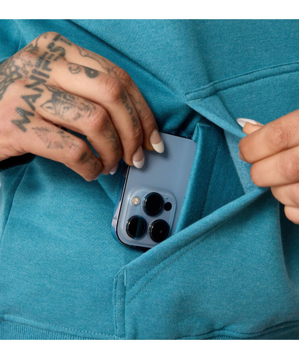 A close-up of a person wearing a teal blue hoodie, showing a tattooed hand reaching into the front pocket to insert a light blue smartphone.