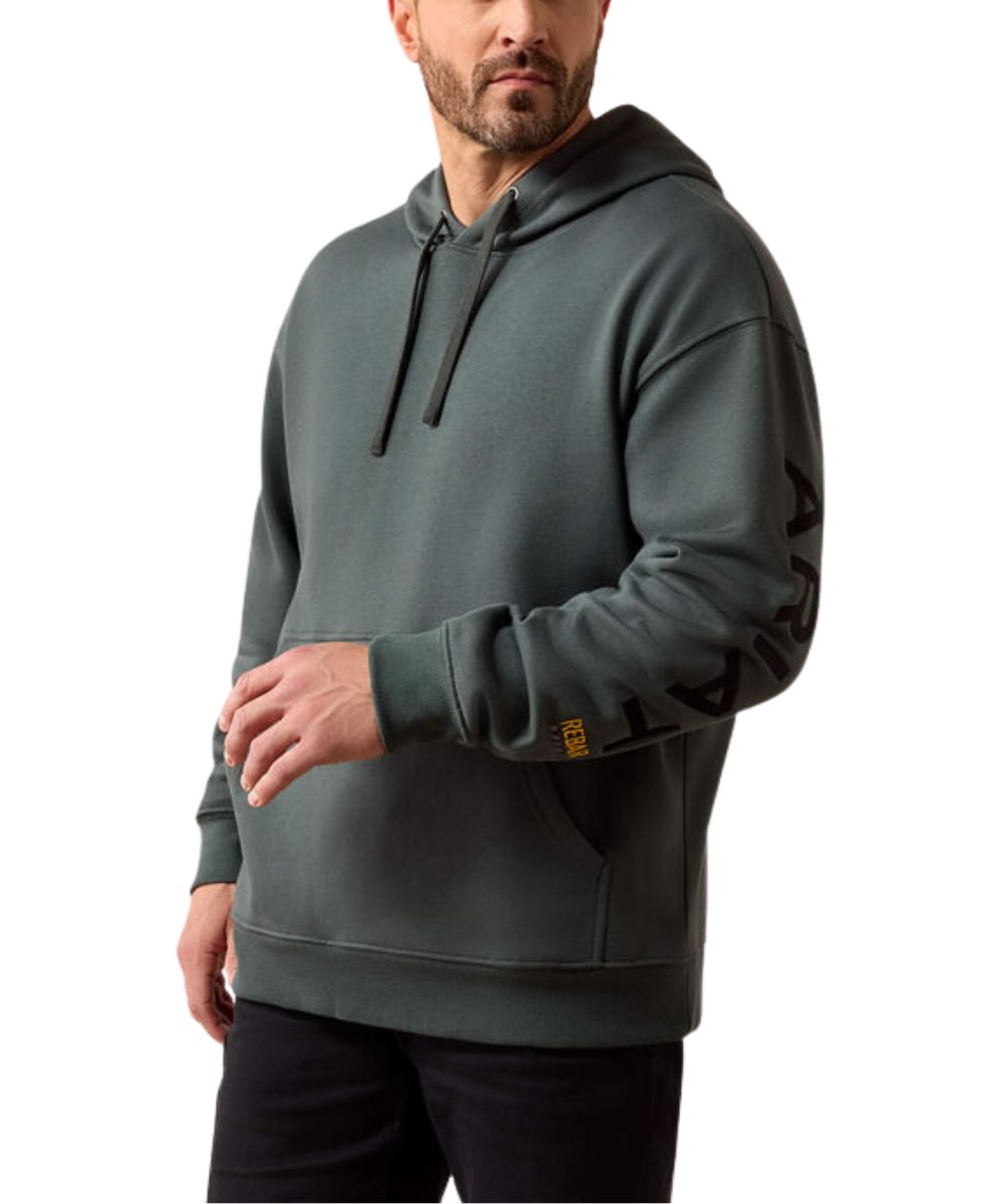 A man wearing a dark gray-green hooded sweatshirt with black drawstrings and a black graphic print on the left sleeve, paired with black pants, against a plain white background.
