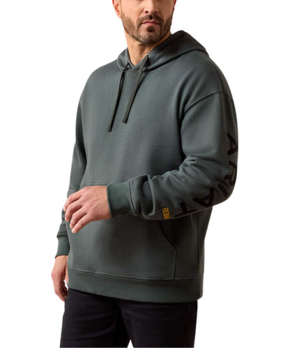A man wearing a dark gray-green hooded sweatshirt with black drawstrings and a black graphic print on the left sleeve, paired with black pants, against a plain white background.