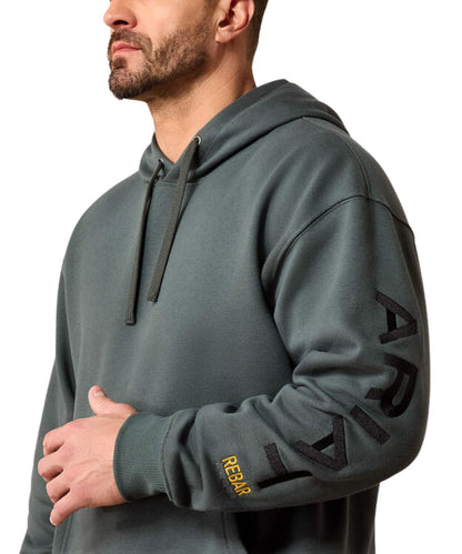 A man wearing a dark gray hoodie with black 'ARIAT' text printed vertically on the right sleeve and a small yellow 'REBAR' logo on the left cuff, shown against a plain white background.
