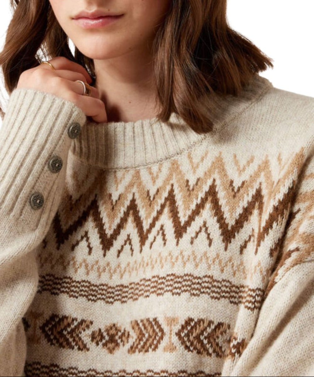 A close-up of a woman wearing a cream-colored knit sweater with brown and beige geometric patterns and faux buttoned cuffs.