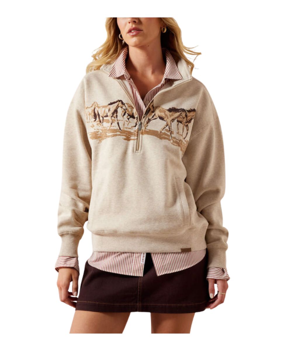 A woman models a beige quarter-zip pullover sweater featuring a graphic print of horses across the chest, layered over a pink and white striped collared shirt and a dark brown mini skirt, against a plain white background.