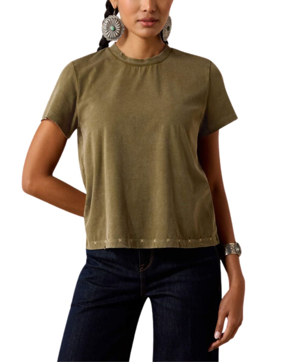 A woman wearing a short-sleeve crewneck t-shirt in olive green with silver studs along the hem, paired with dark blue jeans.