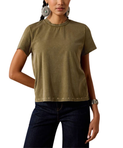 A woman wearing a short-sleeve crewneck t-shirt in olive green with silver studs along the hem, paired with dark blue jeans.