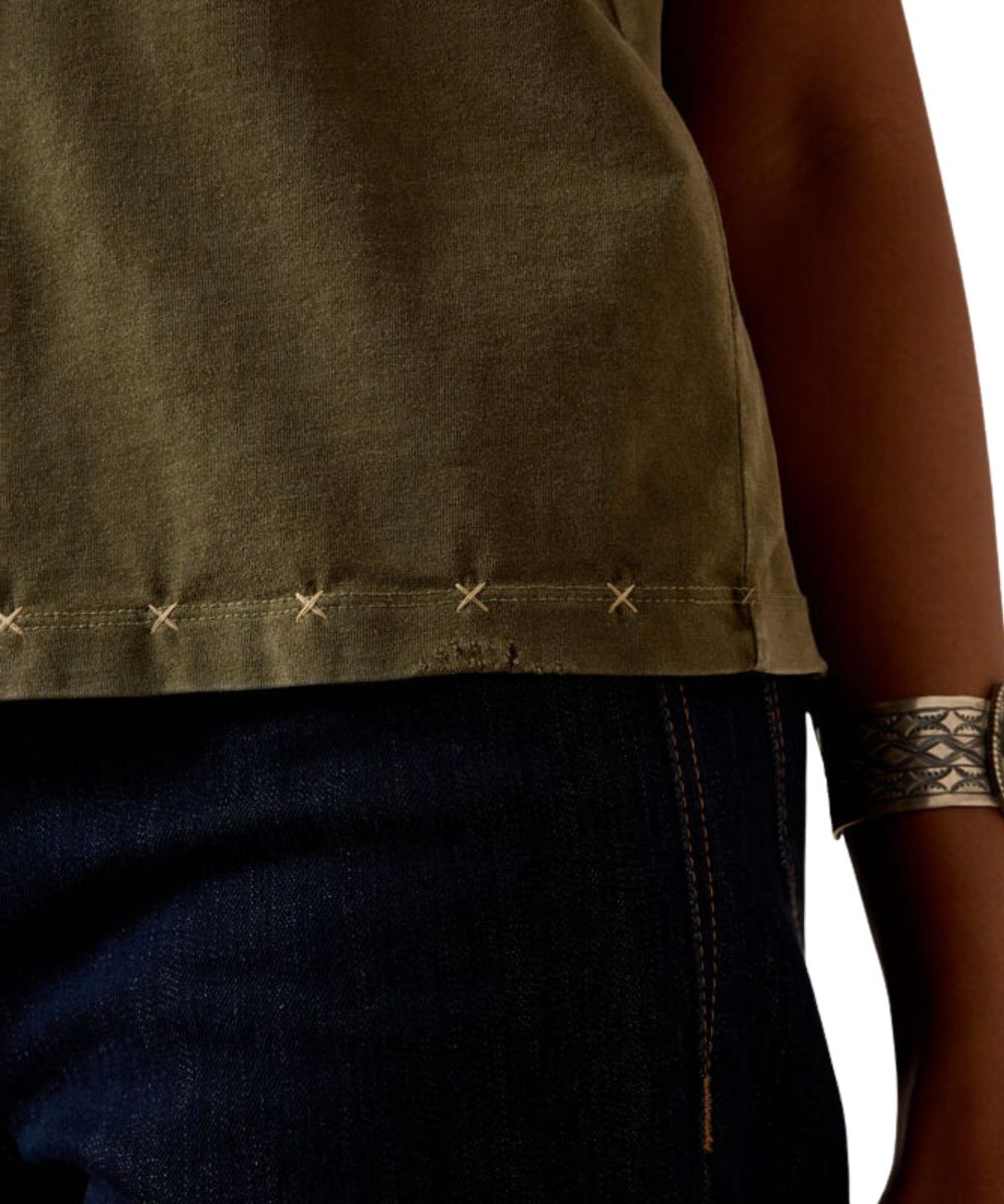 Close-up of a person wearing an olive green sleeveless top with decorative stitching along the hem, paired with dark blue jeans and a patterned metallic cuff bracelet.