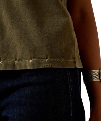 Close-up of a person wearing an olive green sleeveless top with decorative stitching along the hem, paired with dark blue jeans and a patterned metallic cuff bracelet.