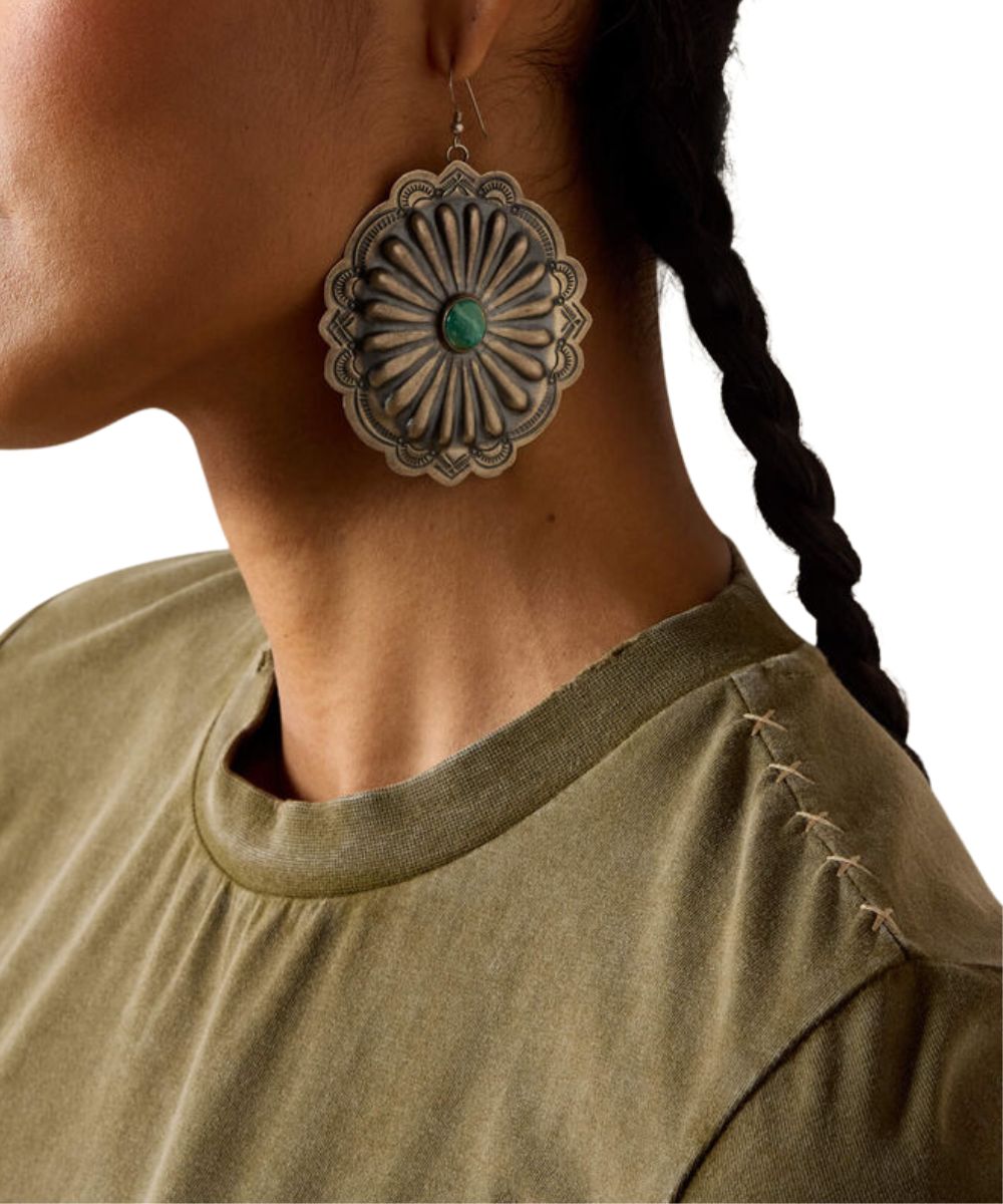 A close-up of a person wearing a large, circular, scalloped silver-toned earring with a green stone in the center. The person is wearing an olive green shirt and has dark hair in a braid against a plain white background.