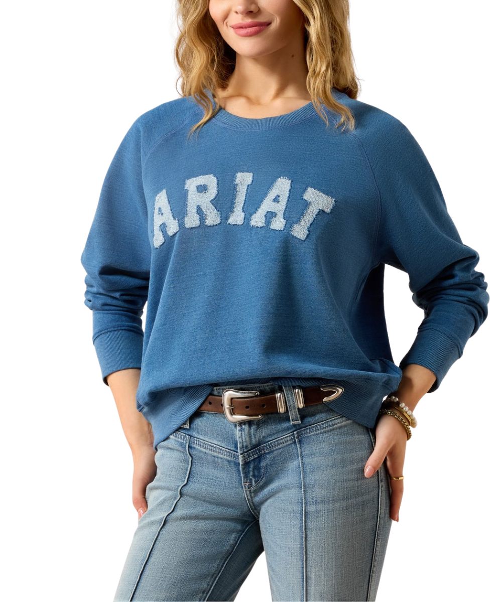 A woman wearing a blue long-sleeve crewneck sweatshirt with 'ARIAT' printed across the chest in white varsity-style letters, paired with light-wash blue jeans and a brown belt, against a plain white background.