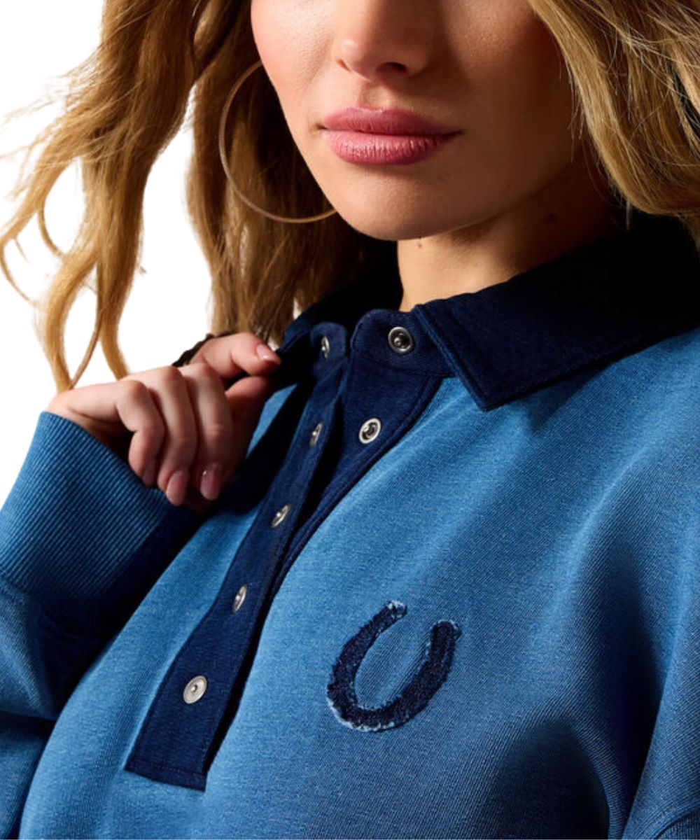 Close-up of a woman wearing a blue knit polo-style top with a dark navy collar and placket, silver buttons, and a dark embroidered horseshoe logo on the chest.