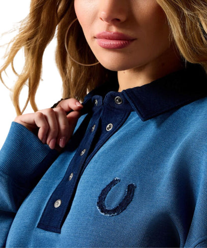 Close-up of a woman wearing a blue knit polo-style top with a dark navy collar and placket, silver buttons, and a dark embroidered horseshoe logo on the chest.