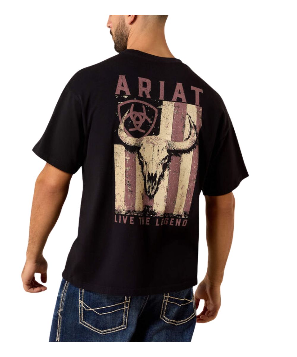 A man wearing a black Ariat t-shirt from the back, featuring a distressed graphic of a skull over a vertical American flag design with the text 'ARIAT' and 'LIVE THE LEGEND'.