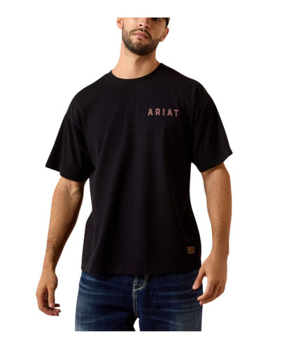 A man wearing a black short-sleeve crewneck t-shirt with 'ARIAT' printed in a dusty rose color on the left chest, paired with blue jeans, against a plain white background.