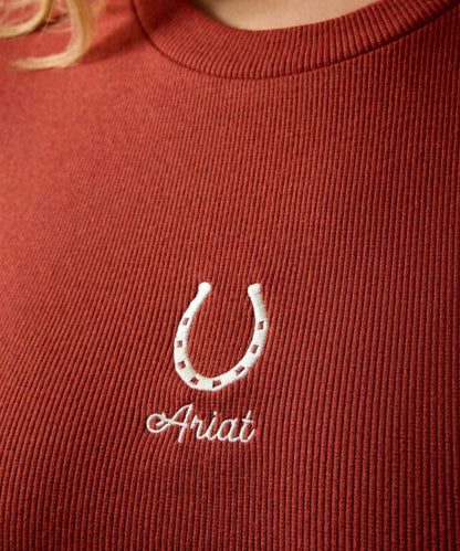 Close-up of a ribbed terracotta red crewneck sweater featuring an embroidered white horseshoe and the name 'Ariat' in white script.
