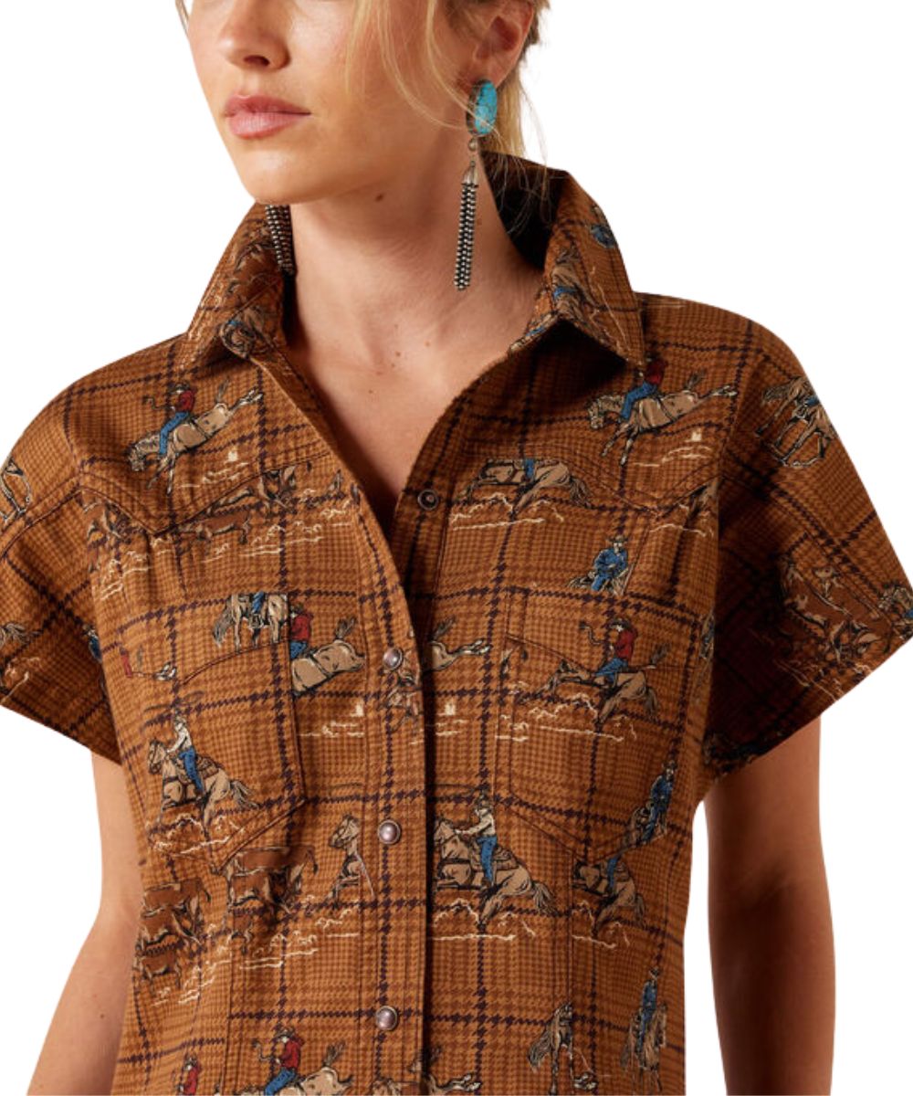 A close-up of a woman wearing a brown short-sleeve button-up shirt with a plaid pattern and a western-style cowboy print, paired with long turquoise dangling earrings against a white background.