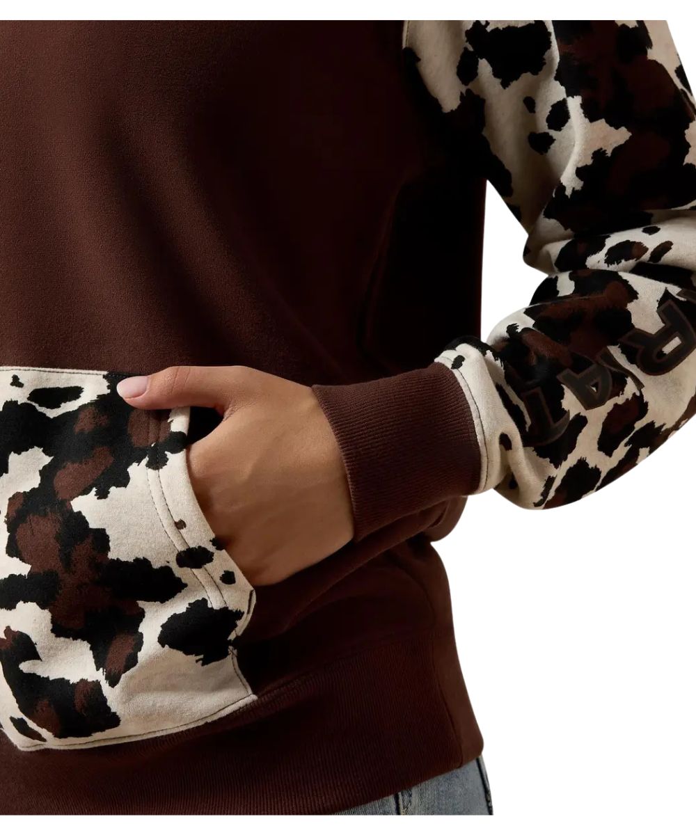 A close-up of a person wearing a brown crewneck sweatshirt with long sleeves featuring a brown, black, and beige cow-print pattern, with a side pocket being accessed by a hand.
