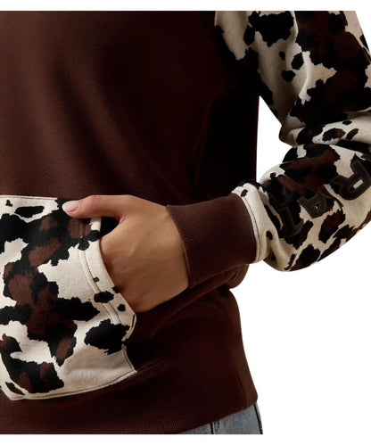 A close-up of a person wearing a brown crewneck sweatshirt with long sleeves featuring a brown, black, and beige cow-print pattern, with a side pocket being accessed by a hand.