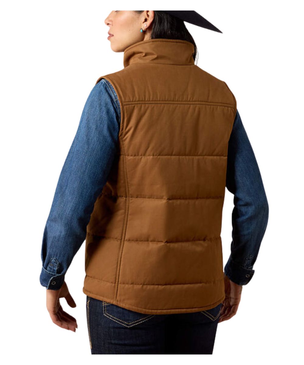 A woman shown from the back wearing a brown quilted puffer vest over a blue denim shirt and dark jeans, against a plain white studio background.
