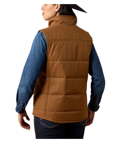 A woman shown from the back wearing a brown quilted puffer vest over a blue denim shirt and dark jeans, against a plain white studio background.