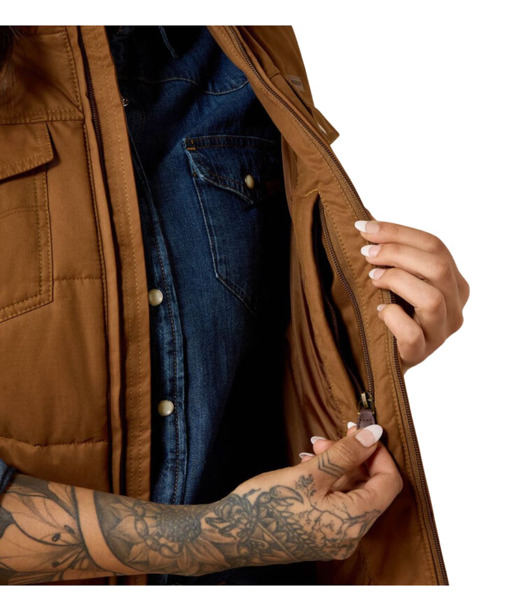 Close-up of a person wearing a brown quilted jacket over a blue denim shirt, with both hands visible as they open the jacket to show a zippered pocket. The background is solid white.
