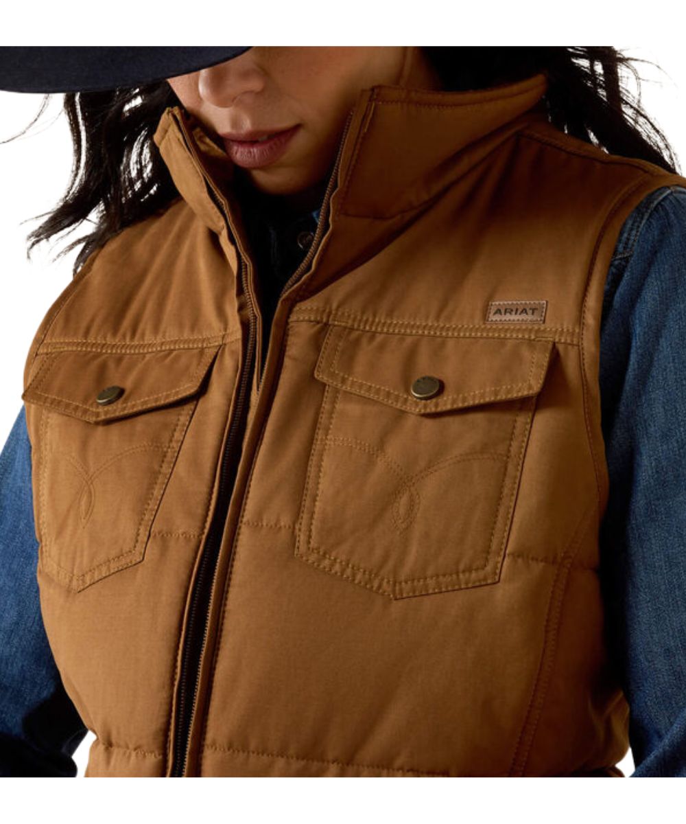 A close-up of a person wearing a brown quilted vest over a blue denim shirt. The vest has a high collar, front zipper, and two chest pockets with button-flap closures.