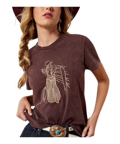 A woman wearing a brown short-sleeve t-shirt with a beige line-art graphic of a cowboy and lasso, paired with dark jeans and a brown hat against a white background.
