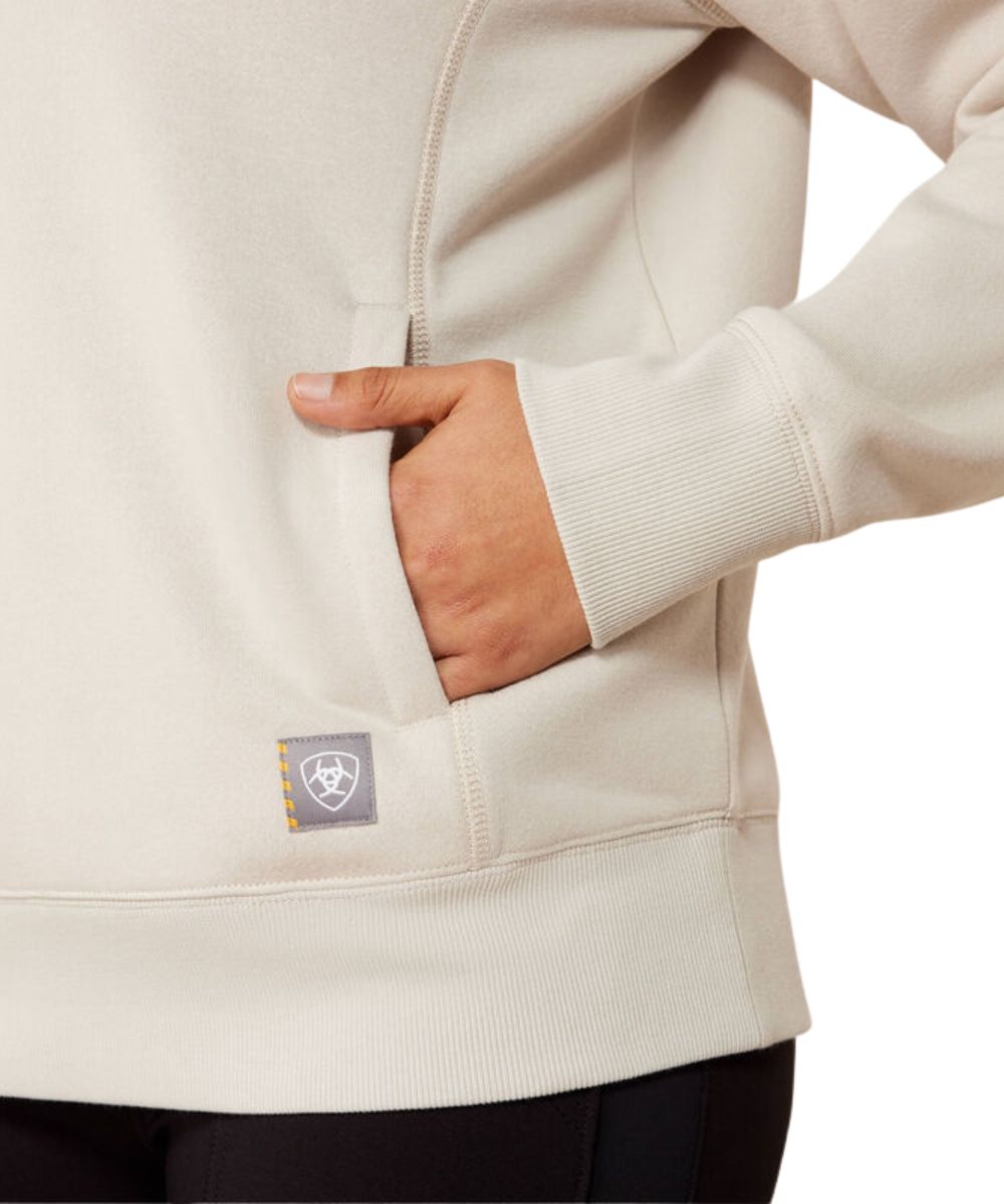 Close-up of a person wearing a light beige fleece hoodie with a side pocket, their hand tucked into the pocket. A small gray brand patch is visible near the pocket.