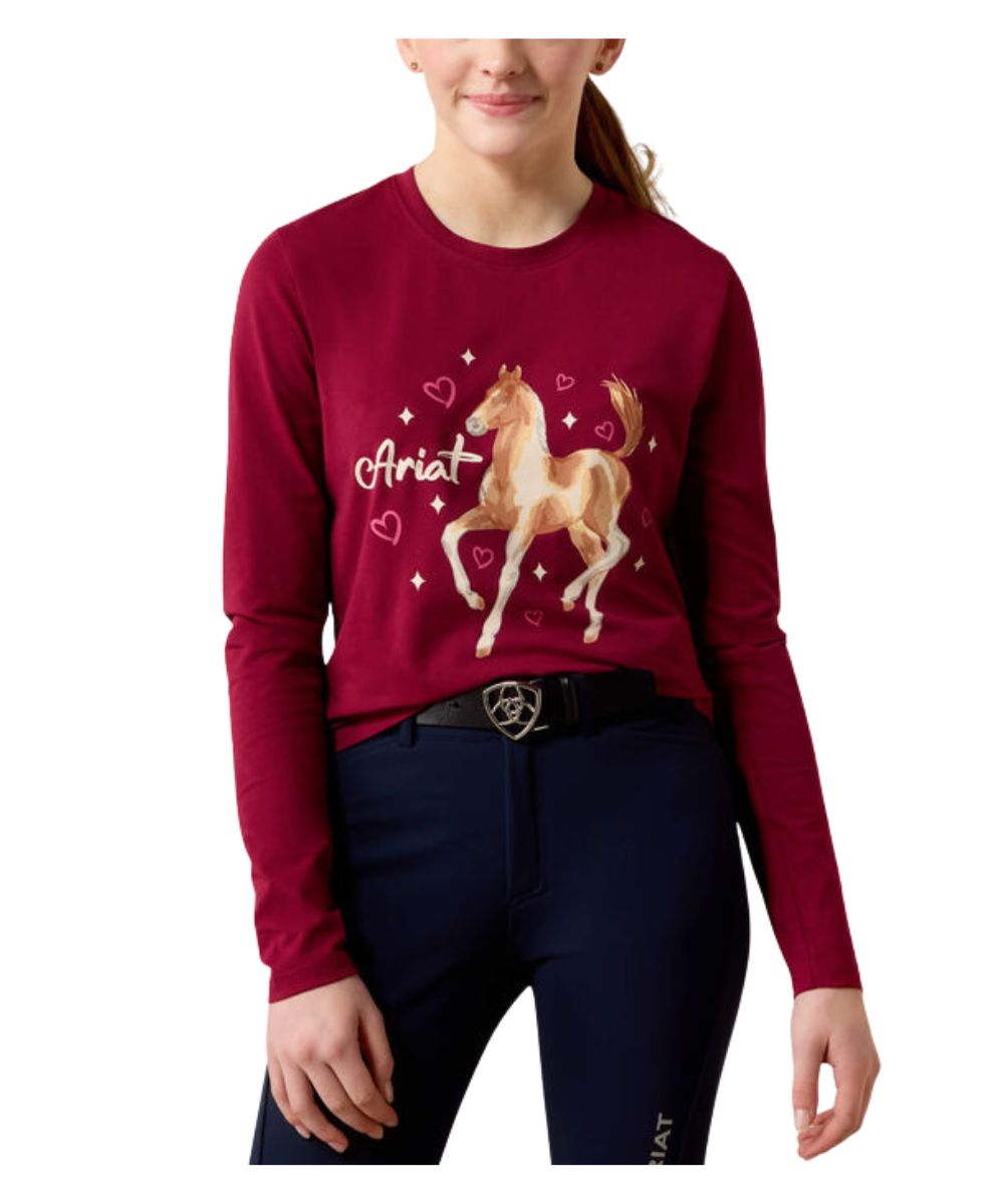 A girl wearing a long-sleeve maroon top with a graphic of a horse and the name 'Ariat' on the front, paired with dark navy riding pants against a white background.