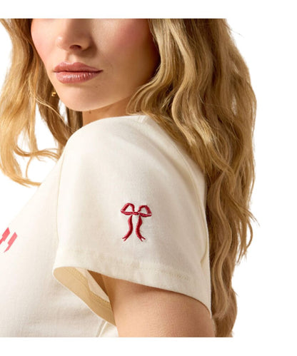 A close-up of a woman wearing a cream-colored short-sleeve t-shirt with a red embroidered bow on the sleeve, shown from the side against a white background.