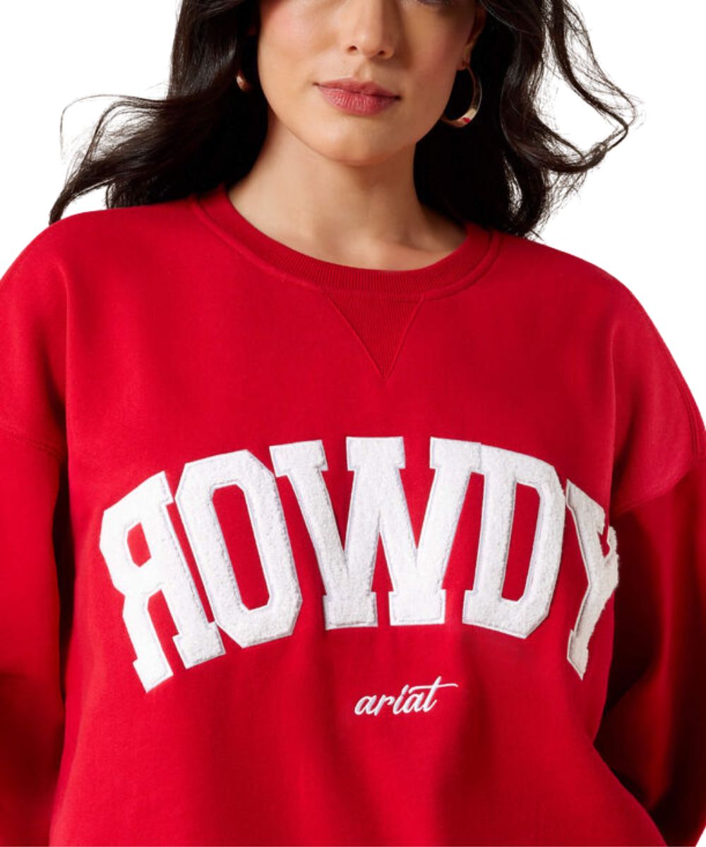 A close-up of a woman wearing a bright red crewneck sweatshirt with 'ROWDY' printed in large white arched letters across the chest and 'ariat' in smaller white script below.