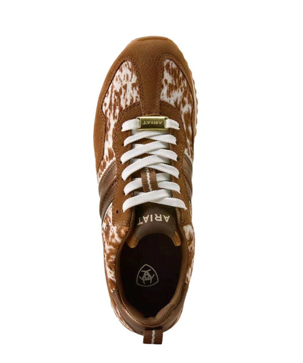 A top-down view of an Ariat sneaker in brown and white cow-print leather with white laces, on a solid white background.