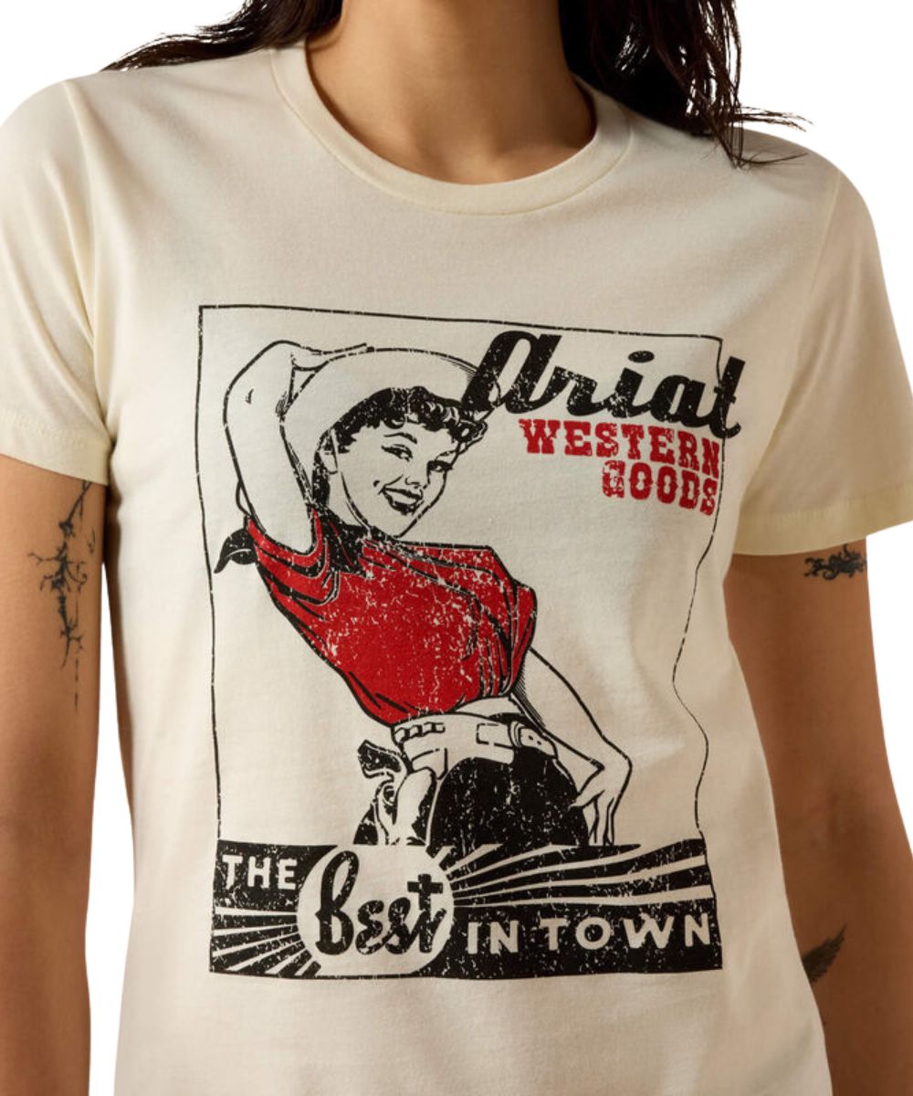 A close-up of a person wearing a cream-colored graphic t-shirt. The shirt features a vintage-style distressed print of a woman in a cowboy hat and red top, with text reading 'Ariat Western Goods' and 'The best in town'.