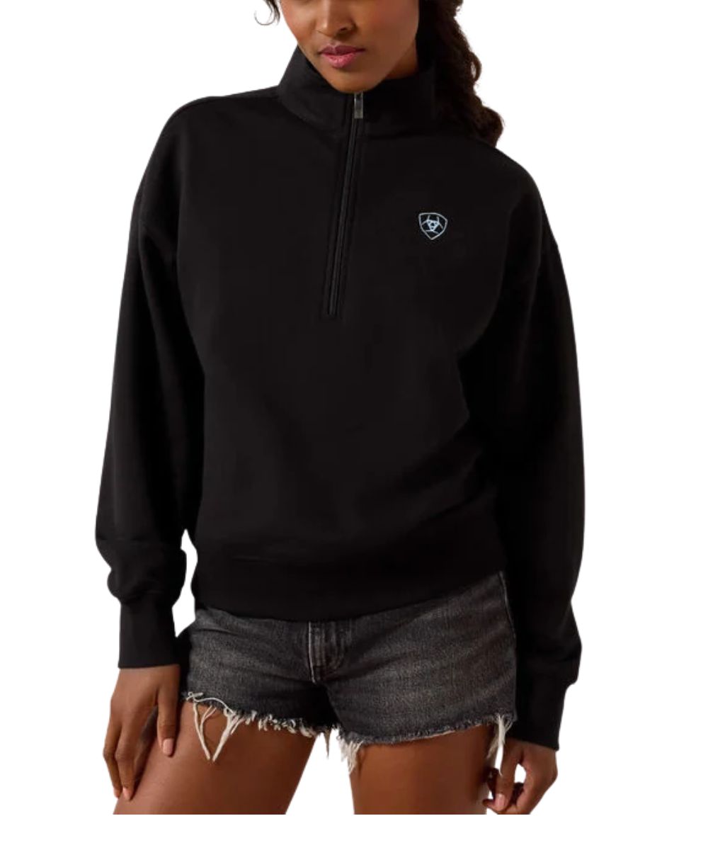 Woman wearing a black half zip sweatshirt with an Ariat logo on the left chest and black shorts