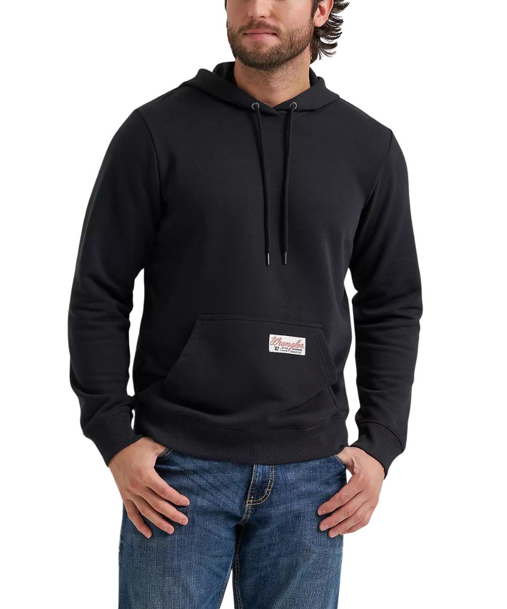 A man wearing a black pullover hoodie with a kangaroo pocket and a small rectangular logo patch on the front, paired with blue jeans against a plain white background.