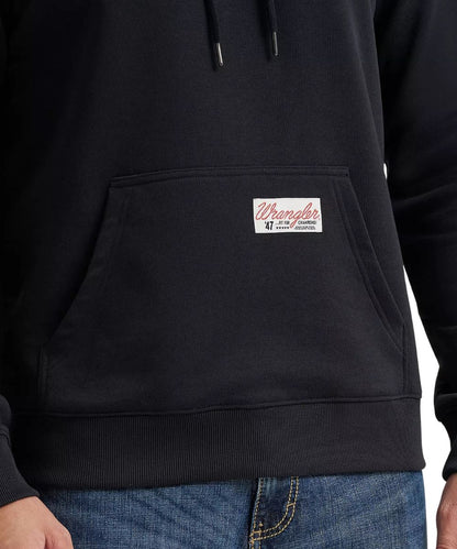 Close-up of a black hoodie worn by a model, showing the front kangaroo pocket with a white Wrangler brand patch and the top of blue jeans.