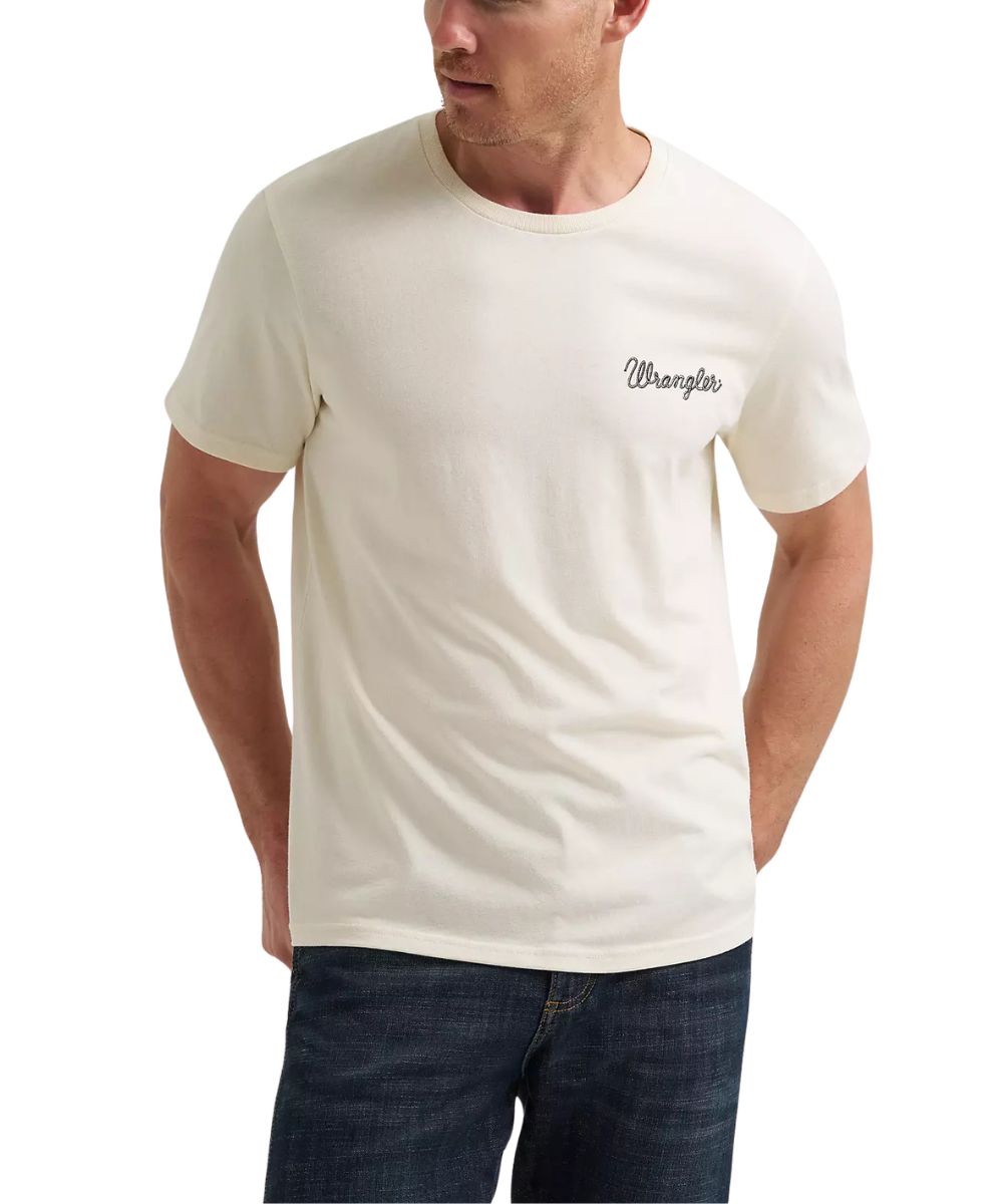 A man wearing a cream-colored Wrangler short-sleeve t-shirt with the brand name printed in script on the left chest, paired with dark blue jeans against a plain white background.