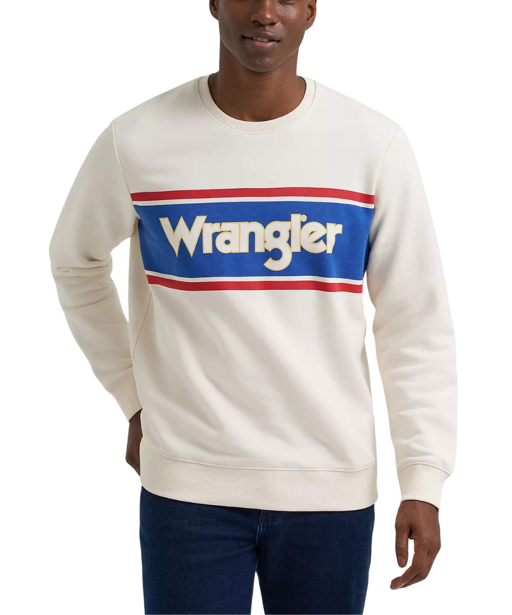 A man wearing a cream-colored Wrangler crewneck sweatshirt with a blue chest graphic featuring red and white stripes and the Wrangler logo, paired with dark blue jeans against a white background.