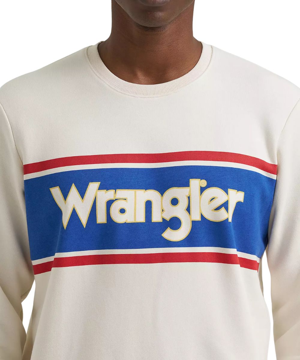 A close-up of a person wearing a Wrangler crewneck sweatshirt. The sweatshirt is off-white with a bold blue chest stripe bordered in red and white, featuring the Wrangler logo in cream lettering.