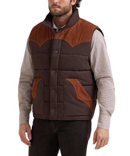 A man wearing a brown quilted vest with contrasting brown suede-like shoulder panels, layered over a beige and white striped long-sleeve shirt and black pants, against a plain white background.